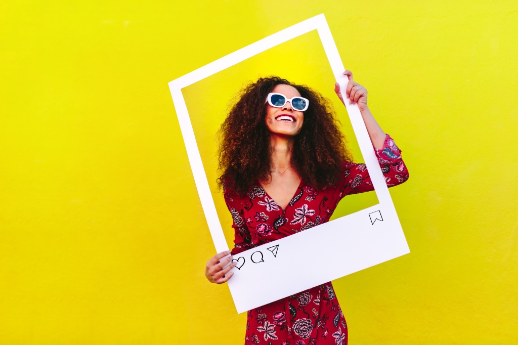 Woman with curly hair wearing sunglasses holding a empty social networking photo frame. Social media influencer standing against yellow looking through at blank photo frame.